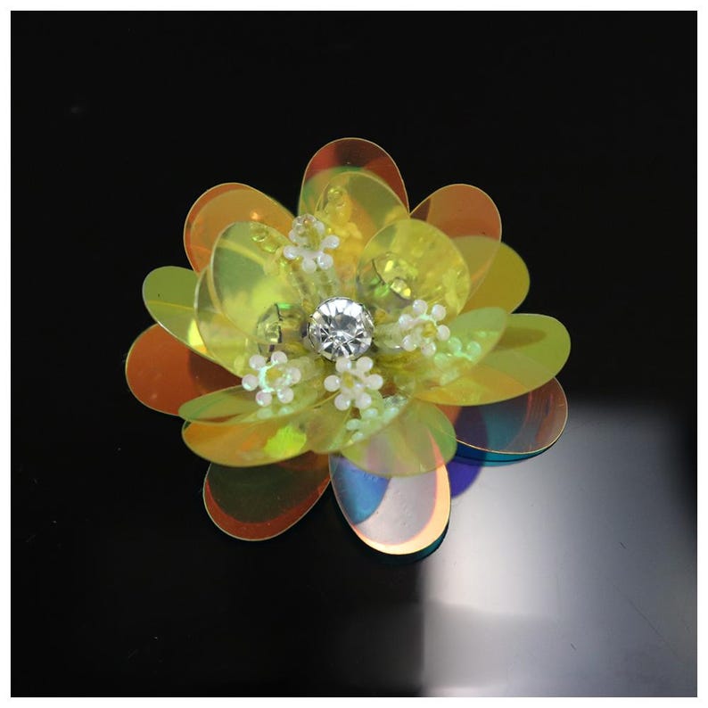 May include: A flower-shaped applique crafted from iridescent yellow and orange sequins. The center features a clear, faceted crystal surrounded by small, white, bead-like floral accents, creating a layered, reflective, and dimensional appearance.