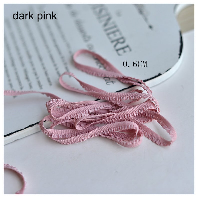 Pu&ograve; includere: Primo piano di un bordo elastico con volant rosa, largo 0,6 cm. Il bordo &egrave; piegato e disposto su una superficie bianca.