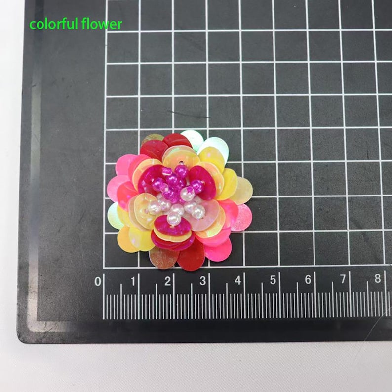 May include: A colorful flower applique crafted from layered sequins in shades of pink, yellow, and white, featuring a center cluster of small purple and white beads. The item is positioned on a black cutting mat with a ruler showing 3 inches.