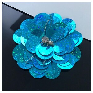 Può includere: Una spilla circolare a forma di fiore realizzata con paillettes turchesi iridescenti a strati. Il centro presenta due strass trasparenti sfaccettati e sottili perline di vetro trasparente sono intervallate tra i petali.