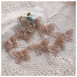 May include: Delicate, light brown butterfly appliques with intricate lace detailing and a pearl bead center. These decorative embellishments are arranged on a white lace surface, perfect for craft projects or adding a touch of elegance.