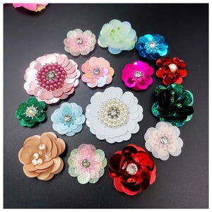Può includere: Una collezione di abbellimenti floreali decorativi in vari colori, tra cui rosa, blu, verde e rosso. Ogni fiore presenta paillettes e centri di perline o strass. Questi materiali per l'artigianato sono ideali per aggiungere stile.