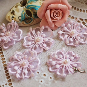 May include: Six pink lace flower embellishments with pearl centers. The flowers are delicate and intricate, with a soft, feminine look.