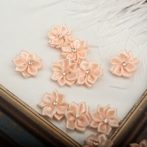 Könnte beinhalten: Pfirsichfarbene Satinbandblumen mit einem Strassstein in der Mitte. Die Blumen sind auf einer weißen Oberfläche angeordnet.