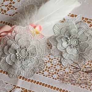 Pode incluir: Dois apliques de flores de renda prateada com um design delicado e intrincado. Os apliques estão sobre uma toalha de renda branca.