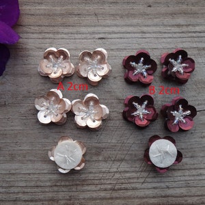 May include: Six flower-shaped sequins with a white center. Two are peach-colored and measure 2 cm. Four are burgundy and measure 2 cm. Two of the burgundy sequins have adhesive backing.