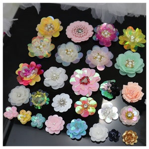 May include: A collection of twenty-five decorative flower appliques in various colors including yellow, pink, white, green, and iridescent tones. Each flower features layered petals and central embellishments made of faux pearls or crystals.