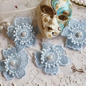 Peut inclure: Quatre appliques de fleurs en dentelle bleu clair avec des centres de perles. Les appliques sont disposées sur un fond de dentelle blanche.