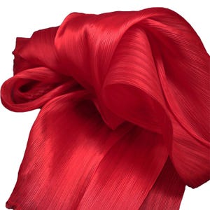 May include: A vibrant red scarf featuring a fine, vertical pleated texture throughout the fabric. The material is lightweight and semi-sheer, draped in soft, flowing folds that create a dynamic, layered appearance with a subtle sheen across the surface.