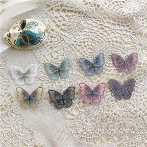 Peut inclure: Assortiment de huit appliques brodées en forme de papillon de différentes couleurs, notamment bleu clair, rose, gris et blanc. Les appliques sont disposées sur une surface en crochet blanc.