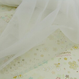 May include: White sheer fabric with a delicate texture. The fabric is draped over a background with a floral pattern and the text "Sweet" and "Hello Remember" in a light pink color.