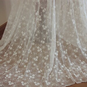May include: White floral lace fabric with a delicate pattern of small flowers and vines. The lace is sheer and has a soft, flowing texture.