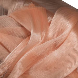 Peut inclure: Un morceau de tissu couleur pêche avec une texture finement plissée. Le matériau est semi-transparent et léger, présentant un motif côtelé vertical cohérent qui crée des reflets et des ombres subtils sur toute la surface pliée.
