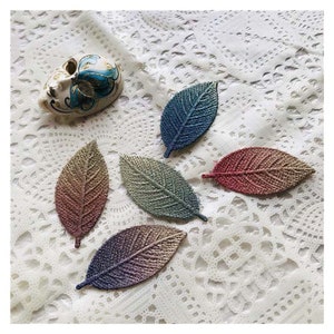 以下が含まれることがあります： 青、緑、ピンク、紫のグラデーションで染められた葉の形をした布のアップリケが5つ。アップリケは白いレースのテーブルクロスの上に並べられています。