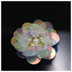 May include: A decorative flower applique crafted from layered, iridescent, semi-transparent circular sequins. The center features a cluster of white faux pearls surrounded by small, gold-toned metal rings embedded with tiny clear crystals.
