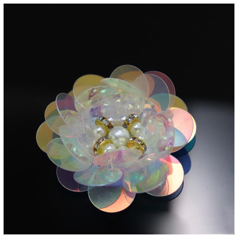 May include: A decorative flower applique crafted from layered, iridescent, semi-transparent circular sequins. The center features a cluster of white faux pearls surrounded by small, gold-toned metal rings embedded with tiny clear crystals.