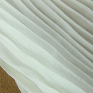 Puede incluir: Tela blanca plisada con una textura suave y fluida. La tela es ligera y transparente, con un brillo sutil.