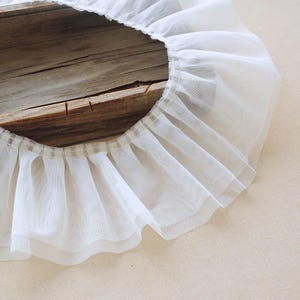 Puede incluir: Primer plano de un ribete de tul blanco fruncido. El ribete está en capas, creando un efecto de volantes. La tela es semitransparente y el ribete está sobre una superficie de madera.
