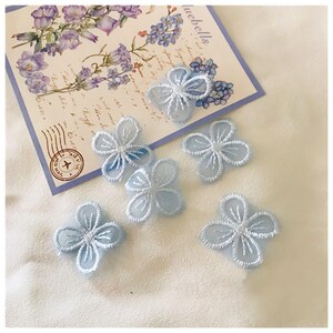 Puede incluir: Seis apliques de encaje de color azul claro con un centro blanco. Los apliques están dispuestos sobre una superficie blanca con una postal que presenta un diseño floral y la palabra "bluebells" en la esquina superior derecha.
