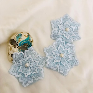 Puede incluir: Tres parches bordados en forma de flor de color azul claro con detalles de hilo blanco y una perla blanca en el centro de cada flor, colocados junto a una máscara decorativa de color crema con detalles pintados en azul y dorado.