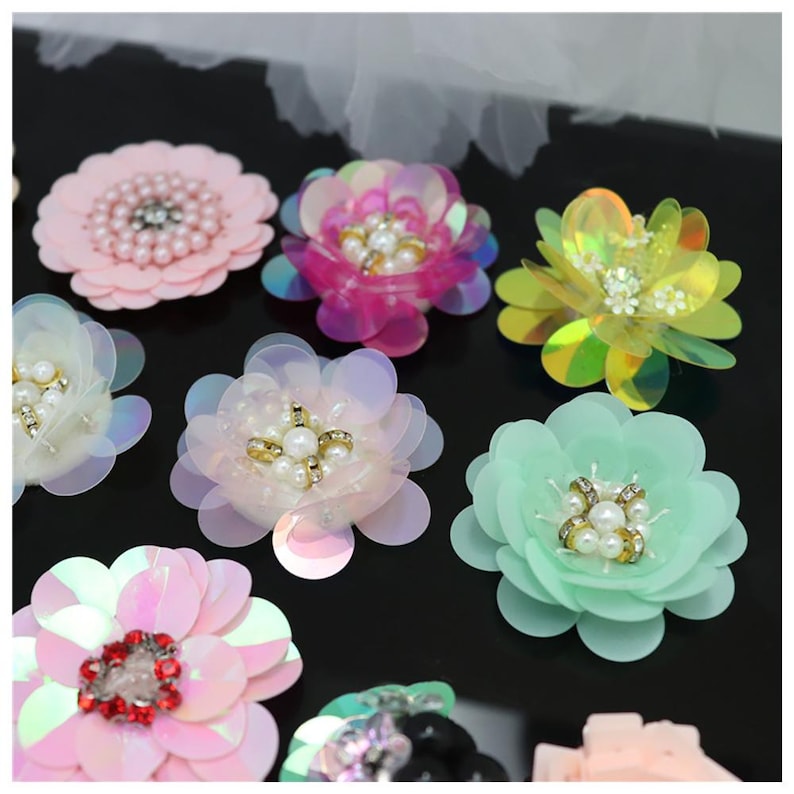 May include: A collection of decorative flower appliques featuring layered petals in various colors including pink, mint green, and iridescent tones. Each flower is centered with a cluster of white faux pearls and small clear rhinestones.