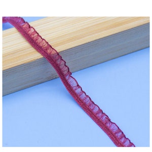 May include: A close-up of a burgundy ruffled trim ribbon. The ribbon is on a wooden surface and is slightly curled.