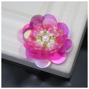 May include: A floral applique featuring layered, iridescent pink sequins arranged in a circular petal pattern. The center of the flower is embellished with a cluster of small white faux pearls and four short, thin bars set with clear crystals.