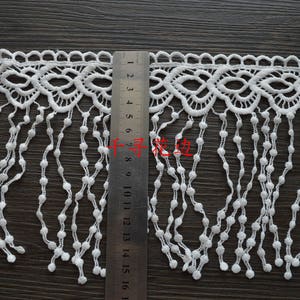 May include: White lace trim with a scalloped edge and a fringe of small white beads. The trim is approximately 16 inches long.