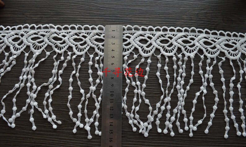 May include: White lace trim with a scalloped edge and a fringe of small white beads. The trim is approximately 16 inches long.