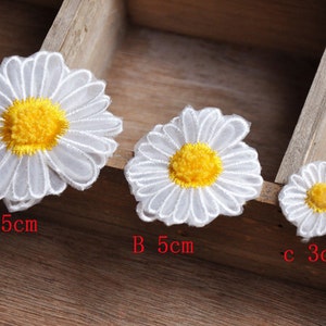 May include: Three white daisy-shaped fabric appliques with yellow centers. The appliques are arranged in a row from largest to smallest. The largest applique is 6.5 inches in diameter, the middle applique is 5 inches in diameter, and the smallest applique is 3 inches in diameter.