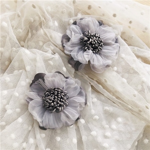 Peut inclure: Deux fleurs en tissu gris avec un centre texturé sur un fond de maille blanche à pois.