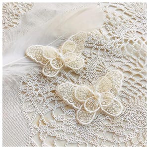 May include: Two cream-colored, lace butterfly appliques with pearl-like centers rest on a cream-colored, open-weave fabric. A white feather is placed near the butterflies. The butterflies have detailed, openwork wings.