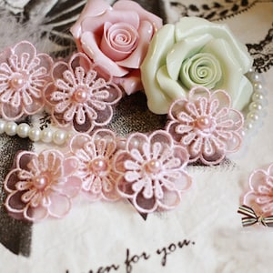 Peut inclure: Des embellissements floraux en dentelle rose avec des centres de perles. Les embellissements sont disposés sur une surface blanche avec un collier de perles et deux fleurs artificielles.