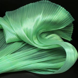 Op de afbeelding: Een groene, iriserende geplisseerde stof met een glinsterende afwerking. De stof is zacht en vloeibaar, met een delicate textuur.