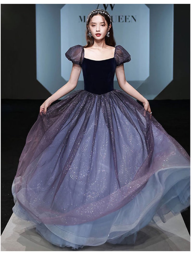 K&ouml;nnte beinhalten: Ein blaues und violettes Ombre-T&uuml;ll-Ballkleid mit einem Samt-Oberteil und Puff&auml;rmeln. Das Kleid hat ein glitzerndes Finish.