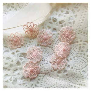 May include: A collection of delicate, blush-pink floral appliques. Each applique features a detailed, openwork design with a central cluster of petals. The appliques are arranged on a white, lace-like surface, with a feather in the background.