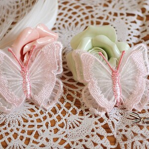 Puede incluir: Dos delicados apliques de mariposa rosa y blanca con un diseño de encaje. Las mariposas están sobre un encaje de ganchillo blanco.