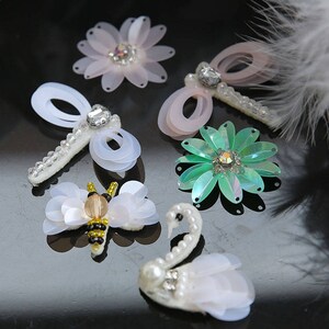 May include: Assortment of colorful fabric appliques featuring a white swan, a bee, a green flower, and two pink dragonflies. The appliques are made of sequins, beads, and fabric.