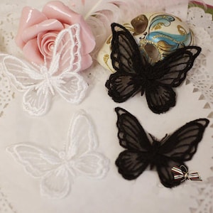Puede incluir: Cuatro delicados apliques de mariposa en encaje blanco y negro. Las mariposas están dispuestas sobre un fondo de encaje blanco.