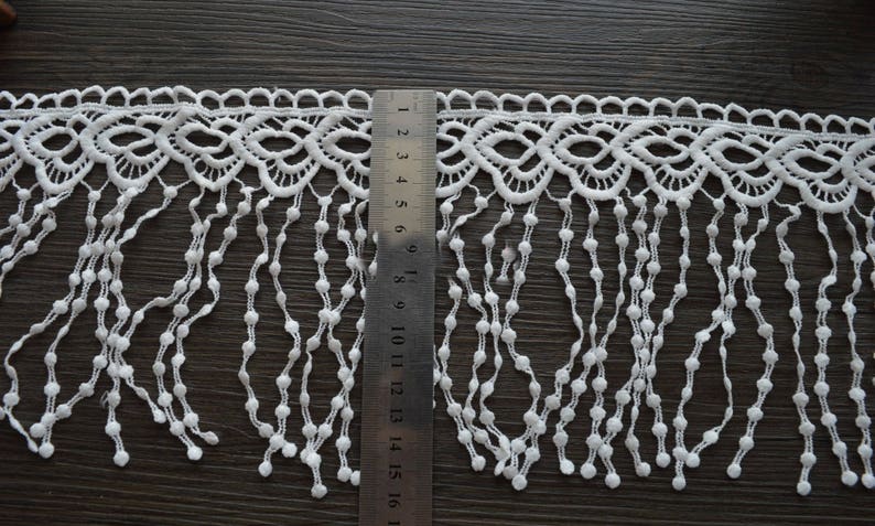 May include: White lace trim with a scalloped edge and fringe. The fringe is made of small white beads and hangs approximately 16 inches long.