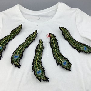 May include: Five peacock feather embroidered patches in black, green and blue colours. The patches are on a white t-shirt.