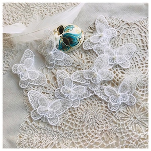 Op de afbeelding: Witte kanten vlinderapplicaties met parelaccenten liggen op een crèmekleurig oppervlak. Een decoratief masker met turquoise en gouden details en een witte veer zijn ook aanwezig. Geschikt voor knutselprojecten.