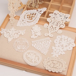Könnte beinhalten: Sortiment von 10 weißen Spitzenapplikationen in verschiedenen Formen und Größen, darunter florale, geometrische und tierische Designs. Die Applikationen sind auf einem beigefarbenen Stoffhintergrund angeordnet.