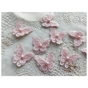 Puede incluir: Una colección de delicadas aplicaciones de mariposas de encaje rosa claro. Cada mariposa presenta detalles intrincados y una pequeña perla redonda en el centro. Las mariposas están dispuestas sobre una superficie texturizada blanquecina.