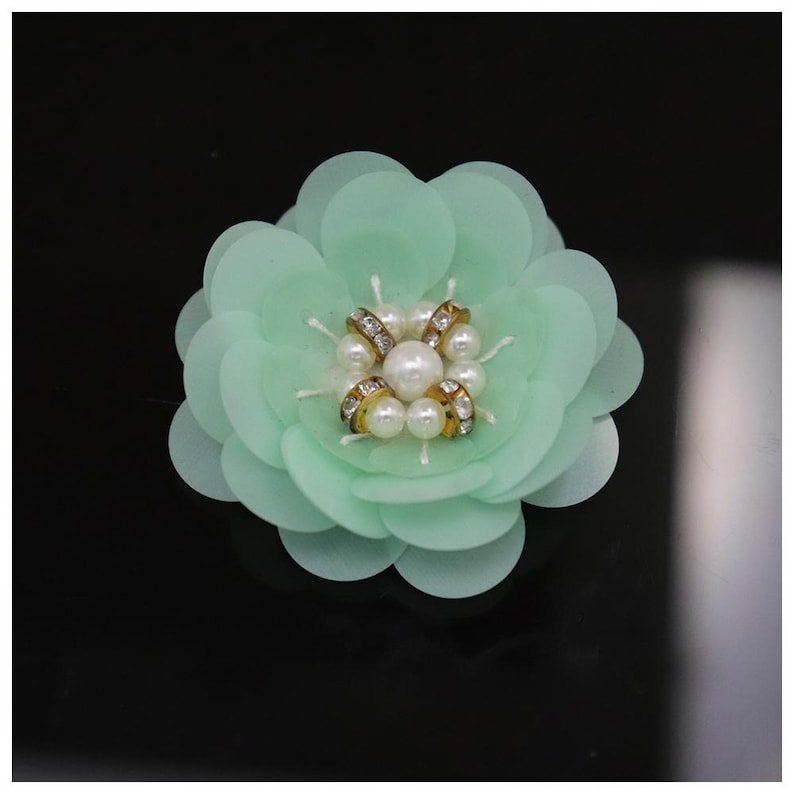 May include: A mint green flower-shaped embellishment featuring layered circular petals. The center is decorated with a cluster of white faux pearls and small gold-toned rings embedded with tiny clear crystals, creating a dimensional floral design.