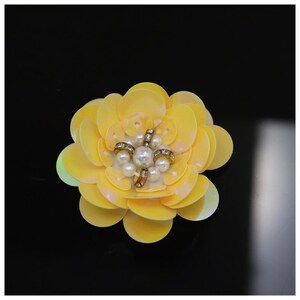 May include: A decorative flower applique crafted from layered, pale yellow circular sequins. The center of the flower features a cluster of small white faux pearls arranged around a central pearl, accented by four small metallic, crystal-set rings.