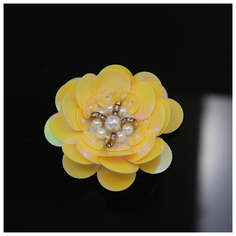 May include: A decorative flower applique crafted from layered, pale yellow circular sequins. The center of the flower features a cluster of small white faux pearls arranged around a central pearl, accented by four small metallic, crystal-set rings.