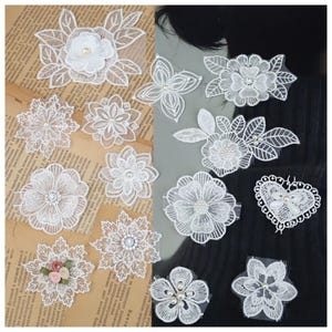 Pode incluir: Uma coleção de acessórios de cabelo de renda branca com vários desenhos florais. O sortido inclui clipes, grampos e apliques em forma de flor, alguns com enfeites de pérolas e strass. Os detalhes delicados da renda criam uma estética romântica e feminina.