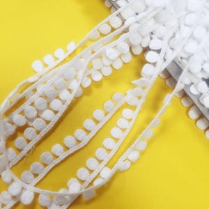 May include: White cotton trim with small round pom-poms. The trim is folded in half and laid on a yellow surface.