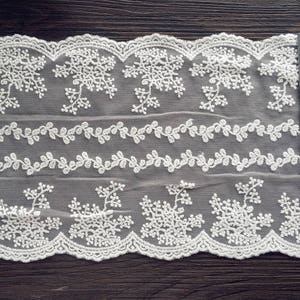May include: Off-white lace fabric with floral and leaf patterns. The lace features a scalloped edge and is measured with a ruler marked in inches. The fabric is likely used for sewing, crafts, or decorative purposes.